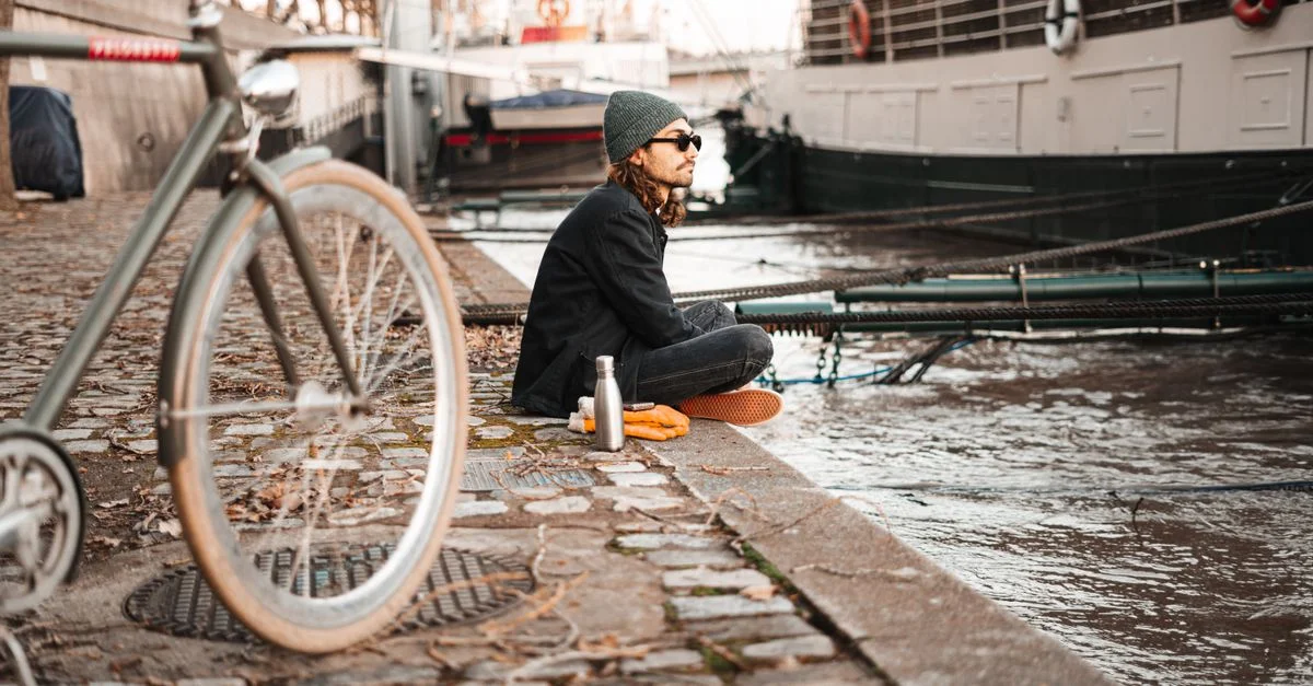 Jeune homme assis au bord d'une rivière avec un vélo, incarnant le mode de vie urbain et la détente.