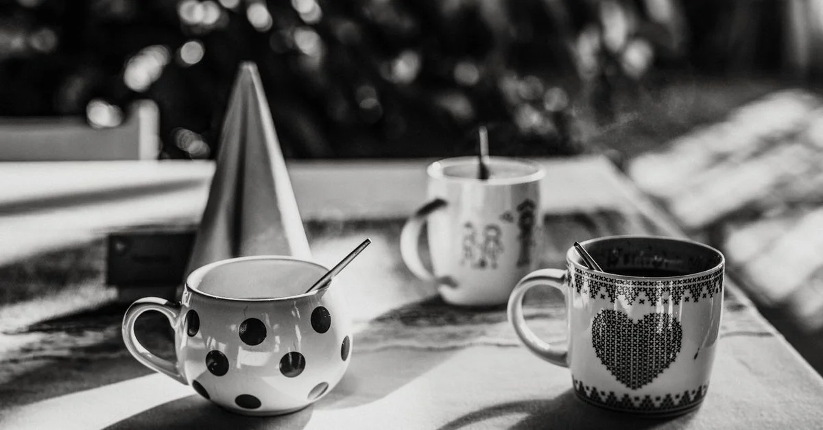 Image en noir et blanc de tasses à café avec cuillères sur une table ensoleillée créant des ombres artistiques.