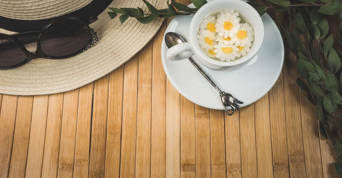 Vue de dessus d'un élégant service de thé avec une tasse de fleurs de camomille, un chapeau et des lunettes de soleil sur un tapis en bambou.