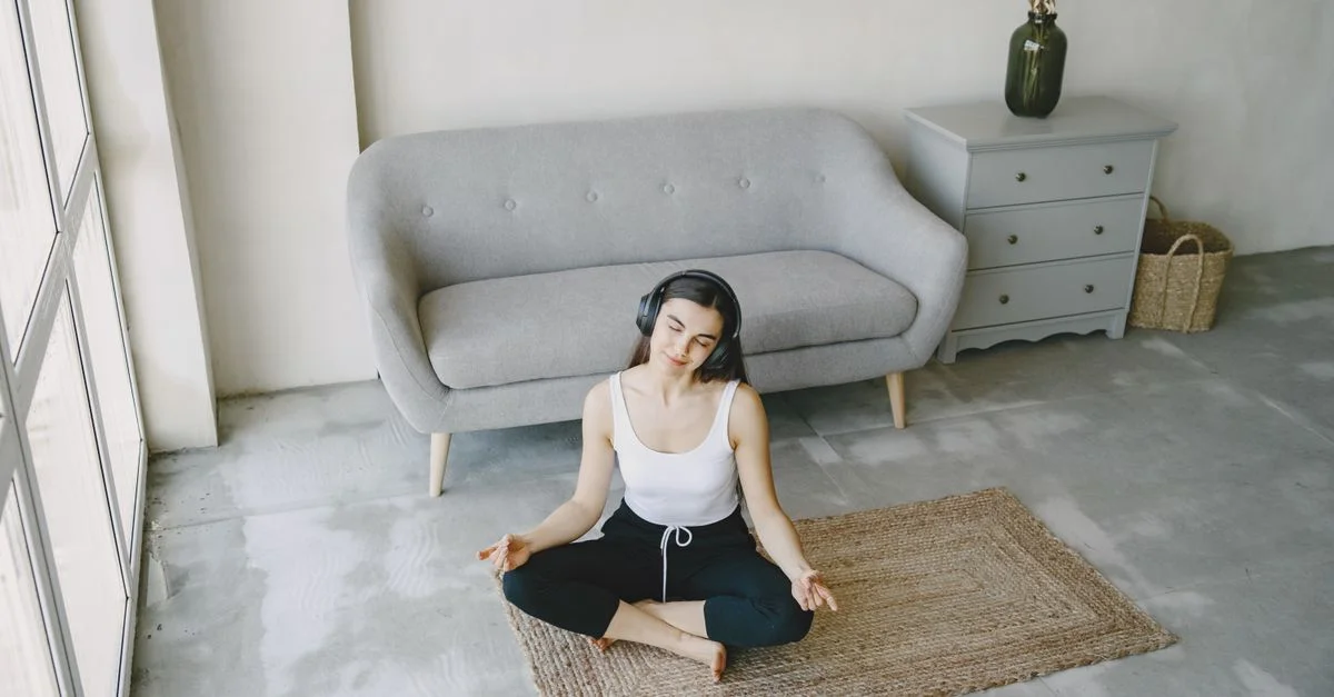 Une jeune femme est assise sur un tapis dans un salon contemporain, méditant avec des écouteurs.