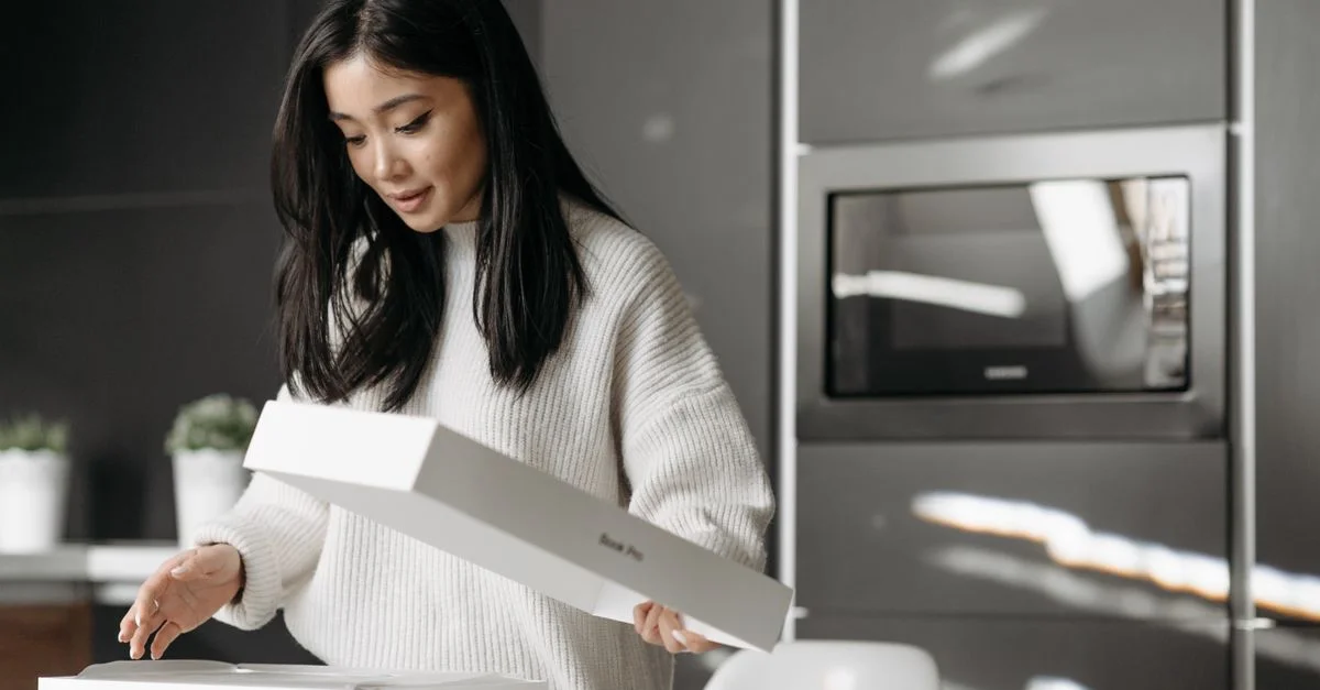 Une femme asiatique déballant avec enthousiasme une boîte de livraison dans sa cuisine moderne.