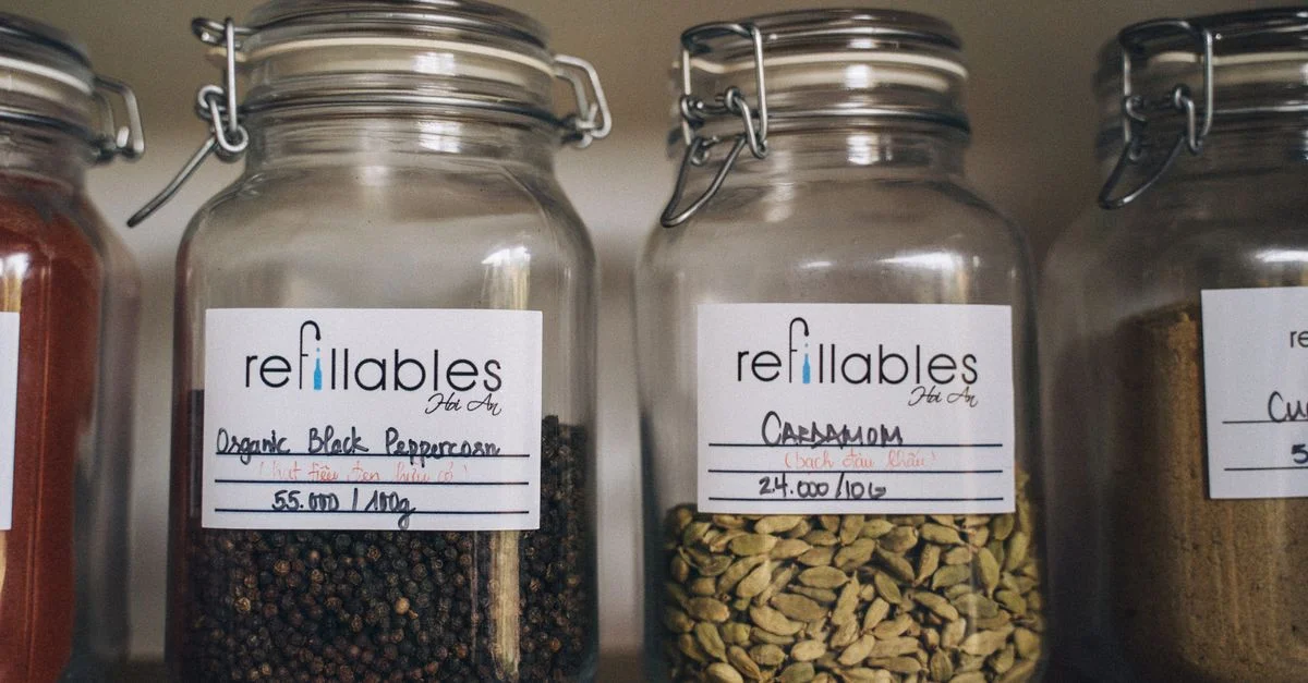 Une collection d'épices dans des bocaux en verre rechargeables et écologiques sur une étagère en bois, promouvant un stockage alimentaire durable.